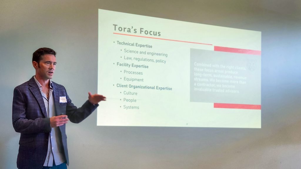 Brandon Mogan, Tora President, presenting at a conference in front of a projected presentation slide about Tora's technical, facility, and organizational expertise in environmental compliance.